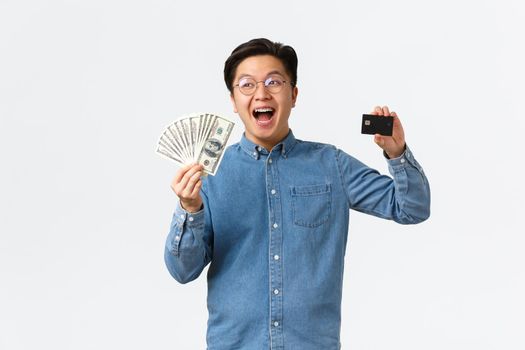 Happy smiling asian man with braces and glasses, laughing cheerfully and showing credit card, holding money, buying something, place cash on bank account, standing white background.