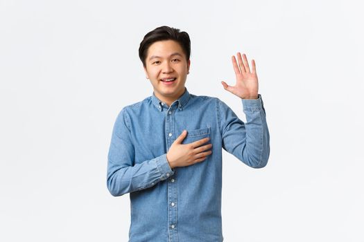 Smiling friendly-looking asian man being honest, raising arm and hold hand over heart while telling truth, making promise or swear something, standing white background sincere.