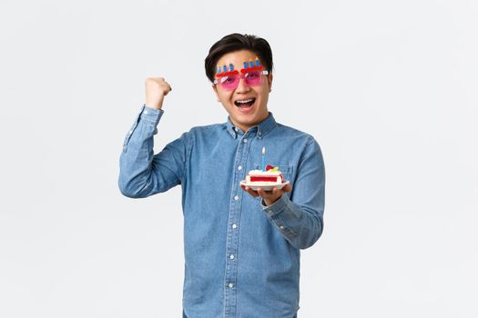 Celebration, holidays and lifestyle concept. Upbeat positive asian guy in funny party sunglasses holding birthday cake and fist pump in hooray gesture, determined bday wish come true.