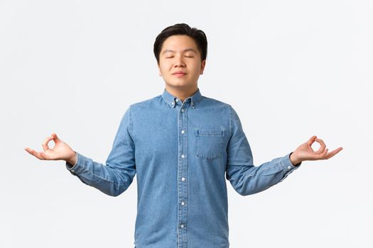 Relieved and relaxed smiling asian man with closed eyes meditating, feeling peaceful and happy, release stress, standing over white background with hands spread sideways in nirvana pose.