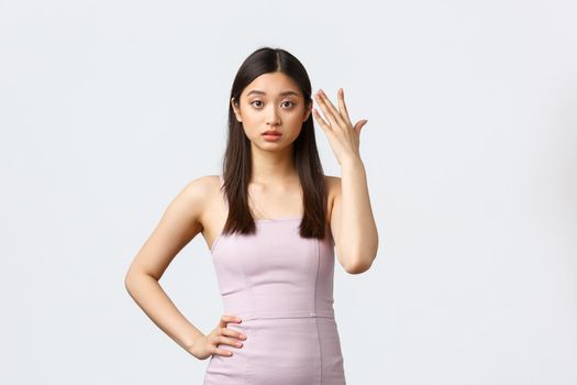 Luxury women, party and holidays concept. Annoyed young asian woman in evening dress, pointing at head and looking irritated, fed up listening complain, standing white background.
