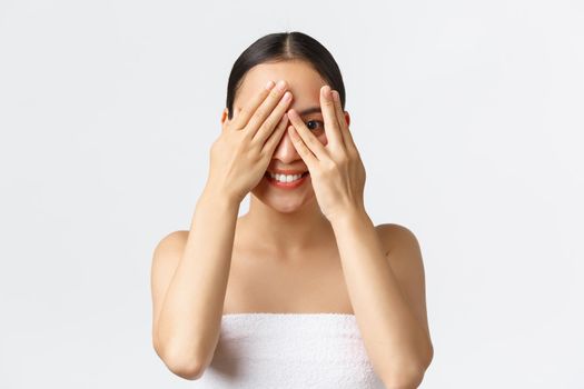 Beauty, cosmetology and spa salon concept. Silly beautiful asian girl in bath towel peeking through fingers as cover eyes with hands and smiling, interested what surprise she except, white background.