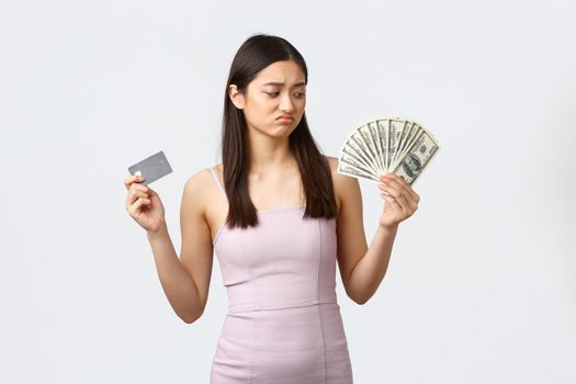 Luxury women, party and holidays concept. Disappointed upset asian girl looking gloomy at money while holding credit card, prefer paying contactless, white background.