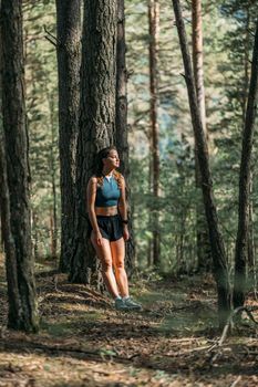 Tired young woman in sportswear leaned on a tree and resting in the forest after working out. Trail running and active life concept