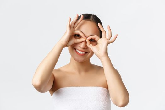 Beauty, cosmetology and spa salon concept. Close-up of playful pretty asian woman in bath towel smiling broadly and making fake glasses with hands over eyes, looking upbeat at camera.