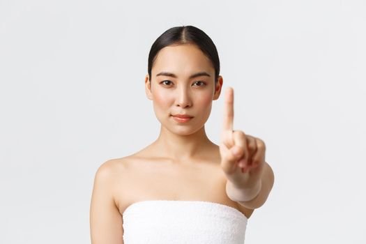 Beauty, cosmetology and spa salon concept. Confident beautiful asian woman showing finger in stop gesture while standing in bath towel, scolding for treating skin bad, forbid action.