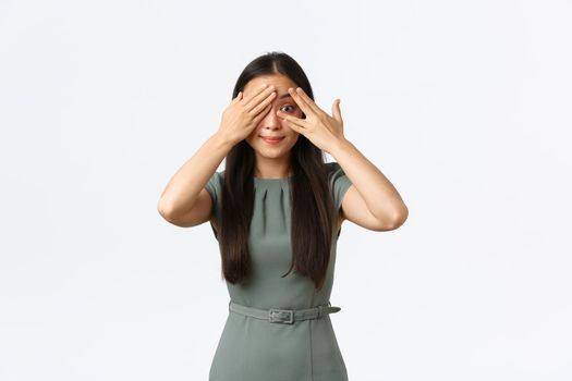 Small business owners, women entrepreneurs concept. Excited asian female in dress, smiling intrigued as shut eyes and peeking through fingers, anticipating surprise, standing white background.