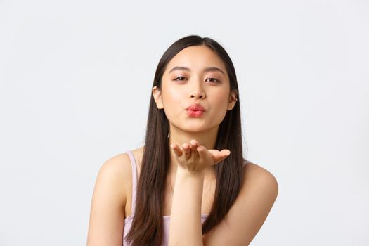 Beauty, fashion and people emotions concept. Close-up portrait of romantic, coquettish beautiful girl in dress, sending air kiss at camera with palm, standing white background.