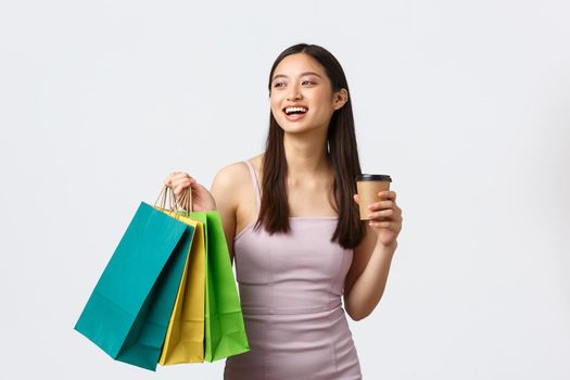 Lifestyle, shopping and tourism concept. Carefree young elegant woman in dress, enjoying weekend, drinking coffee on her way to next shop, carry bags with new clothes, white background.