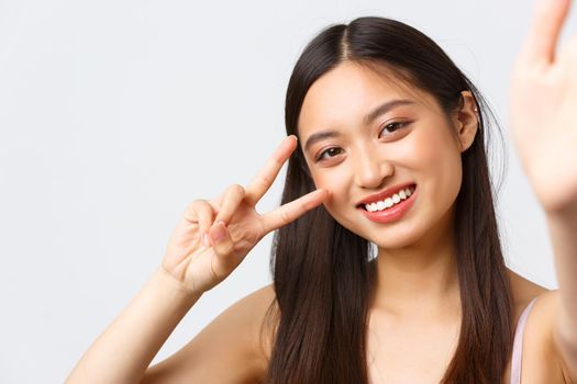 Concept of beauty, fashion and makeup products advertisement. Close-up of beautiful asian female blogger taking selfie or record vlog, showing kawaii peace sign and smiling, extend hand to camera.