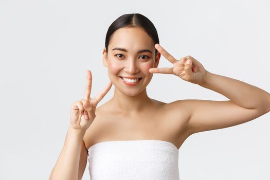 Beauty, cosmetology and spa salon concept. Happy gorgeous asian girl in towel showing kawaii peace gesture and smiling satisfied after beauty clinic, massage therapy, feeling fresh and energized.