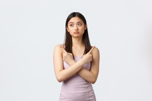 Luxury women, party and holidays concept. Indecisive pretty asian girl in evening dress making tough decision, pointing fingers sideways at both variants, being confused, white background.