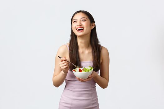 Healthy lifestyle, leisure and people emotions concept. Gorgeous happy asian girl smiling and laughing while eating salad, staying fit for summer, wearing evening dress, white background.