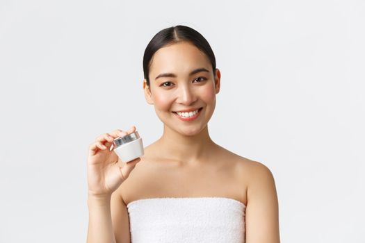 Beauty, personal care, spa salon and skincare concept. Close-up of beautiful asian female in bath towel showing cream and smiling, recommend moisturizing treatment, white background.