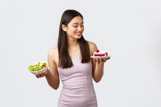 Healthy lifestyle, leisure and food concept. Pleased smiling beautiful asian girl prefer eating sweets instead of fresh salad, looking at piece cake, standing white background.