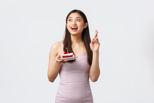 Celebration, party and holidays concept. Hopeful dreamy upbeat birthday girl in dress, cross fingers good luck looking upper left corner, holding b-day cake, white background.