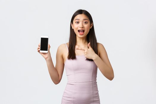 Luxury women, party and holidays concept. Impressed and excited beautiful asian girl in evening dress, talking about application, pointing finger at mobile phone screen, white background.