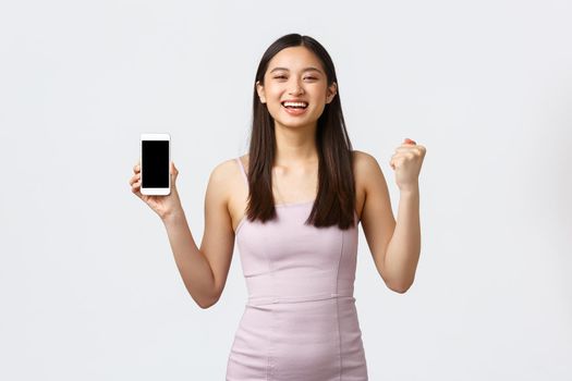 Luxury women, party and holidays concept. Rejoicing, happy asian girl in dress, feeling happy and delighted, got special offer notification on smartphone screen, chanting and show mobile phone.