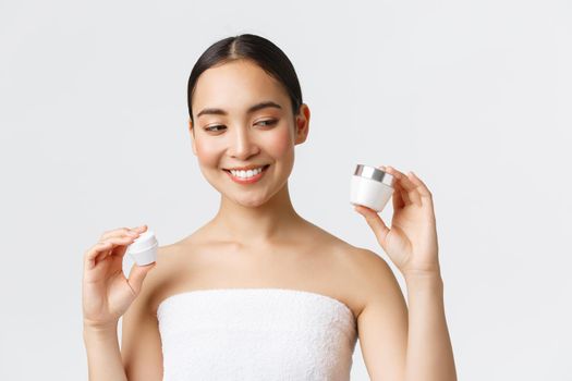 Beauty, personal care, spa salon and skincare concept. Close-up of beautiful asian female in bath towel holding two creams, eye and face nourishing products, smiling, skin treatment.