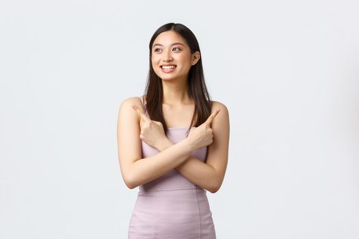 Luxury women, party and holidays concept. Carefree smiling, attractive asian woman in stylish dress, pointing fingers sideways and looking pleased left as made her choice, white background.