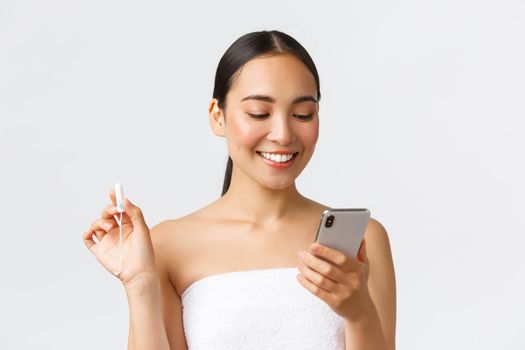Beauty, personal and intimate care, mobile application concept. Smiling pretty asian girl having period, wear towel, looking at menstrual cycle tracker app on smartphone, holding tampon.