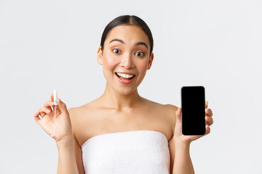 Beauty, personal and intimate care, mobile application concept. Cheerful smiling asian woman showing her menstrual cycle, period tracker on smartphone screen, holding tampon.