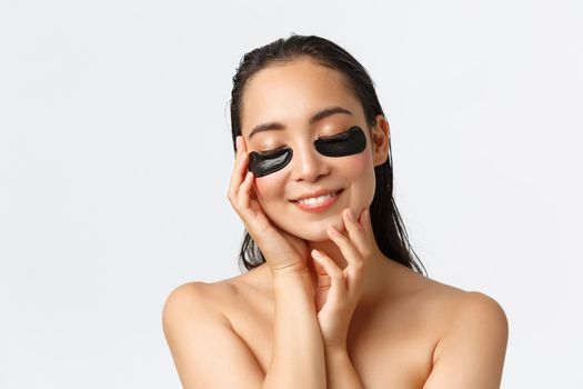 Skincare, women beauty, hygiene and personal care concept. Tender beautiful naked asian woman having spa day, close eyes and smiling as touching skin, apply under eye patches, collagen mask.