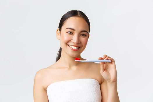 Beauty, personal care, and hygiene concept. Beautiful young asian woman in bath towel smiling white teeth as showing toothbrush, using new toothpaste, recommend product, white background.