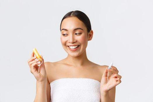 Beauty, personal and intimate care menstrual hygiene concept. Smiling cute asian girl in bath towel holding tampons, looking at one with applicator, making her choice over white background.
