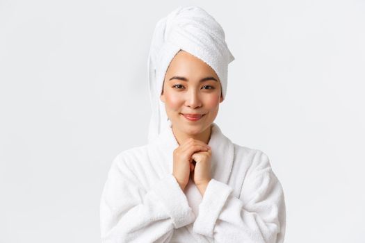 Personal care, women beauty, bath and shower concept. Tender attractive asian girl in bathrobe and towel looking with admiration or delight, smiling cute, standing white background hopeful.