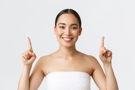 Beauty, cosmetology and spa salon concept. Happy pleased asian female in towel smiling satisfied and pointing fingers up, recommend massage therapy, skincare products, white background.