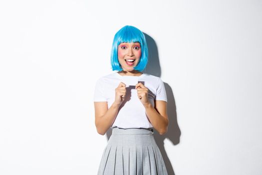 Excited pretty asian girl showing credit card, wearing blue wig for halloween party, standing over white background.