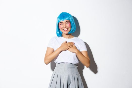 Image of thankful smiling asian girl in halloween costume and blue party wig, holding hands on heart and looking grateful, thanking for something, standing over white background.