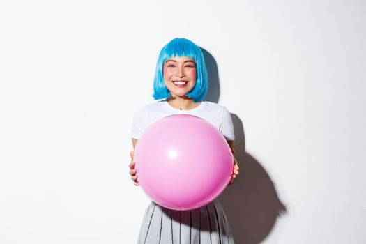 Adorable asian woman celebrating holiday, holding balloon and wearing blue wig for halloween, standing over white background.