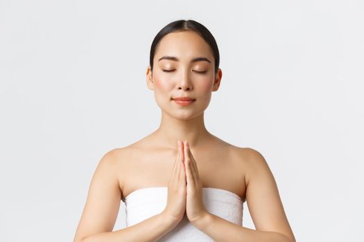 Beauty, cosmetology and spa salon concept. Relaxed sensual pretty asian girl in white towel press hands together and close eyes as meditating, namaste pose while enjoying beauty procedures.