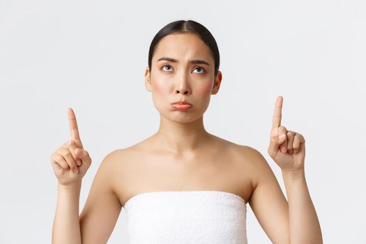 Beauty, cosmetology and spa salon concept. Pouting sad asian girl in white towel sulking and looking, pointing fingers up disappointed, complaining at beauty salon or massage therapy.
