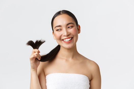 Beauty, hair loss products, shampoo and hair care concept. Gorgeous happy asian woman in bath towel showing hair ends and smiling satisfied, cured split ends and looking satisfied.