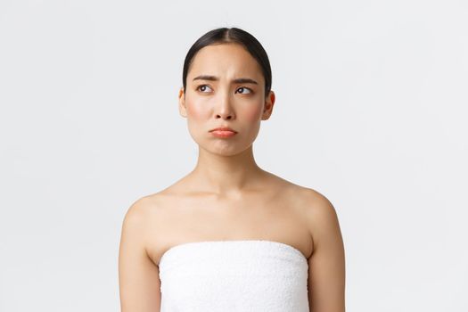 Beauty, cosmetology and spa salon concept. Pouting sad and disappointed asian girl in white towel sulking and looking left with regret, see something upsetting, being jealous.