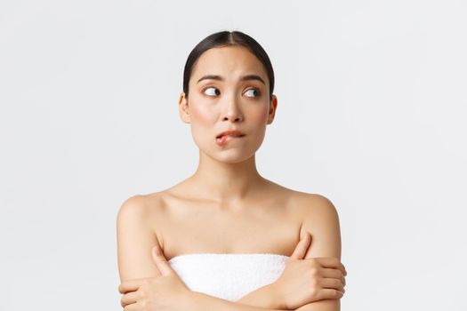 Beauty, cosmetology and spa salon concept. Hesitant and unsure cute asian girl in white towel biting lip and looking away doubtful, worried before depilation procedure in beauty clinic.