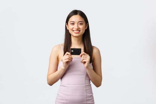 Luxury women, party and holidays concept. Happy rich pretty asian girl in evening dress, showing her brand new credit card, smiling pleased as going shopping, paying for order contactless.