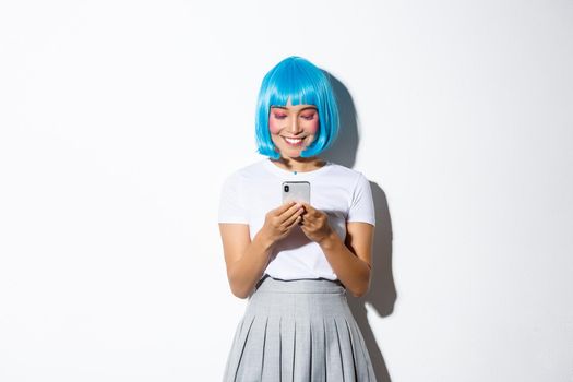 Image of beautiful asian girl looking at mobile phone and smiling, wearing blue anime wig, standing over white background.