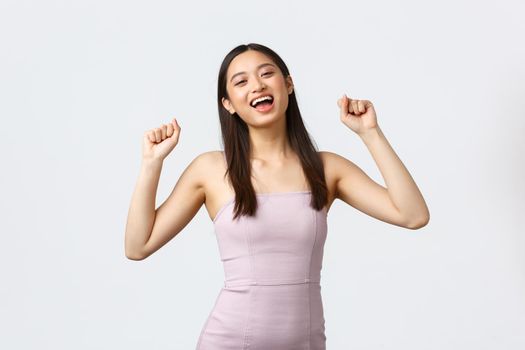Luxury women, party and holidays concept. Relieved and happy pretty korean girl in evening dress, raise hands up shouting yes delighted, winning, become champion, triumphing over white background.