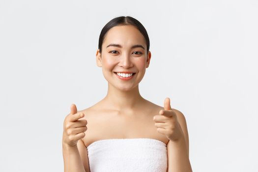 Beauty, cosmetology and spa salon concept. Close-up or attractive asian woman in towel smiling happy and pointing finger at camera, recommend you visit beauty clinic, white background.
