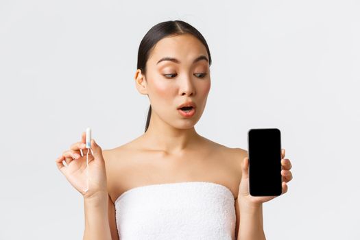 Beauty, personal and intimate care, mobile application concept. Pretty asian woman in bath towel holding tampon and showing smartphone screen with menstrual cycle tracker, period or flow app.