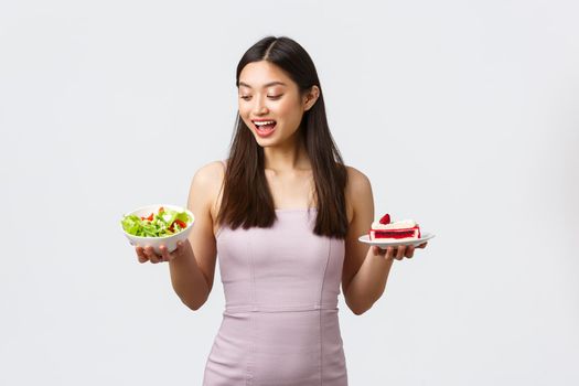 Healthy lifestyle, leisure and food concept. Cheerful beautiful asian girl in dress, looking excited with happy smile at salad instead of sweet cake, standing white background.