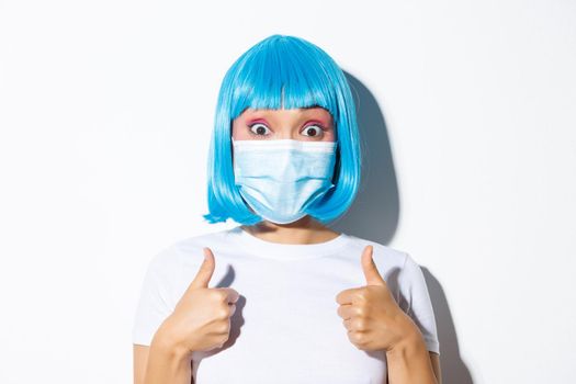 Concept of halloween celebration and coronavirus. Close-up of excited asian girl in medical mask and blue wig showing thumbs-up and approve something good.