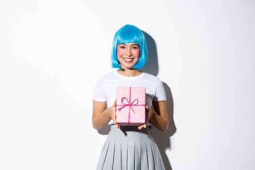 Image of beautiful asian girl in blue party wig, congratulating with holiday, holding gift and smiling, standing over white background.