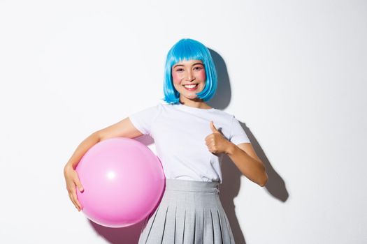 Image of cute asian girl in blue wig and halloween costume, showing thumbs-up, holding large pink balloon.