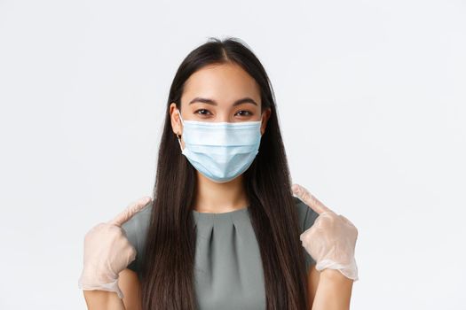 Small business owners, covid-19, preventing virus measures concept. Close-up of smiling asian woman in medical mask and gloves pointing at face, using protective equipment during pandemic.