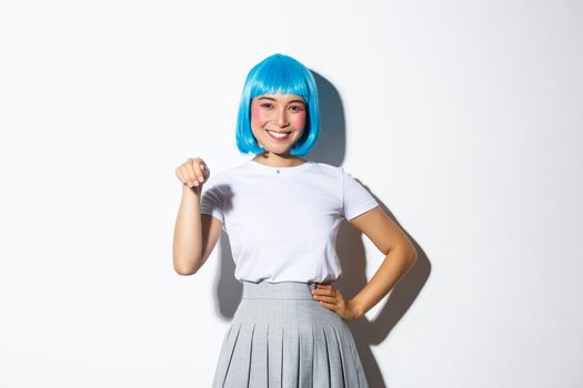 Portrait of lovely asian female in blue party wig, waving hand and smiling, standing happy over white background.
